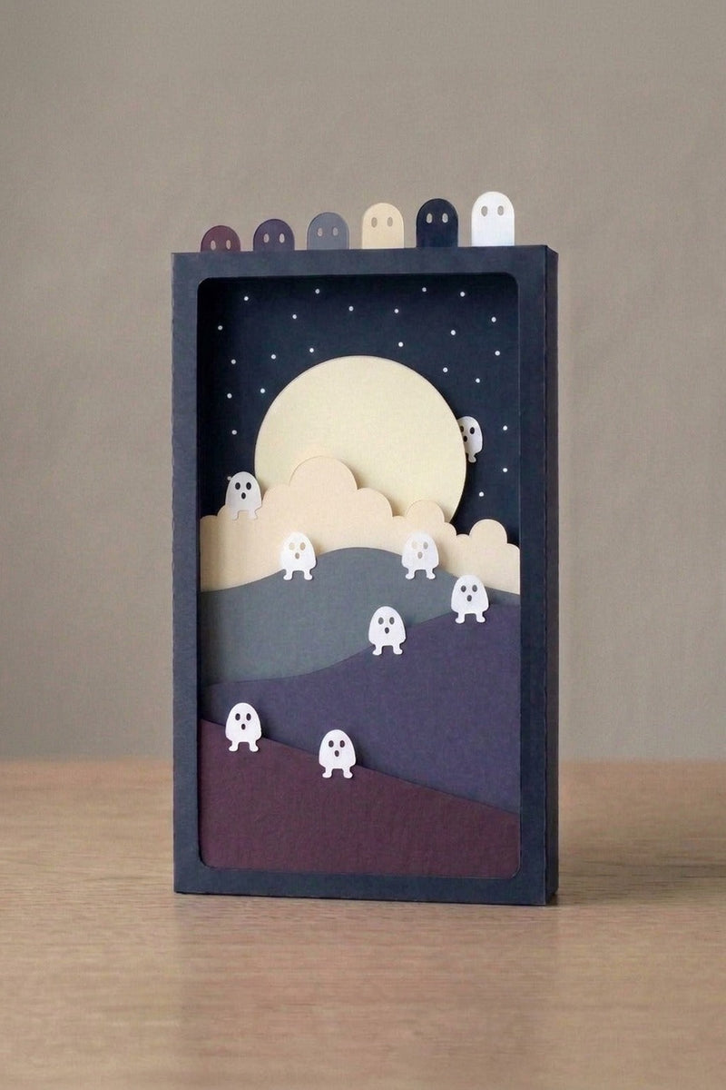 Fluorescent Spirits Pocket Story seen from the front: glowing paper ghosts on violet dunes under a pale moon, in a navy sleeve with six ghost tabs (AI-generated image)