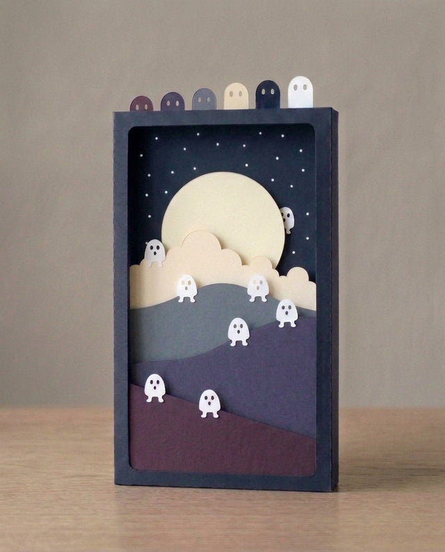 Fluorescent Spirits Pocket Story: eight white paper spirits climb three night hills toward a large ivory moon, layered mass-tinted paper in a navy sleeve