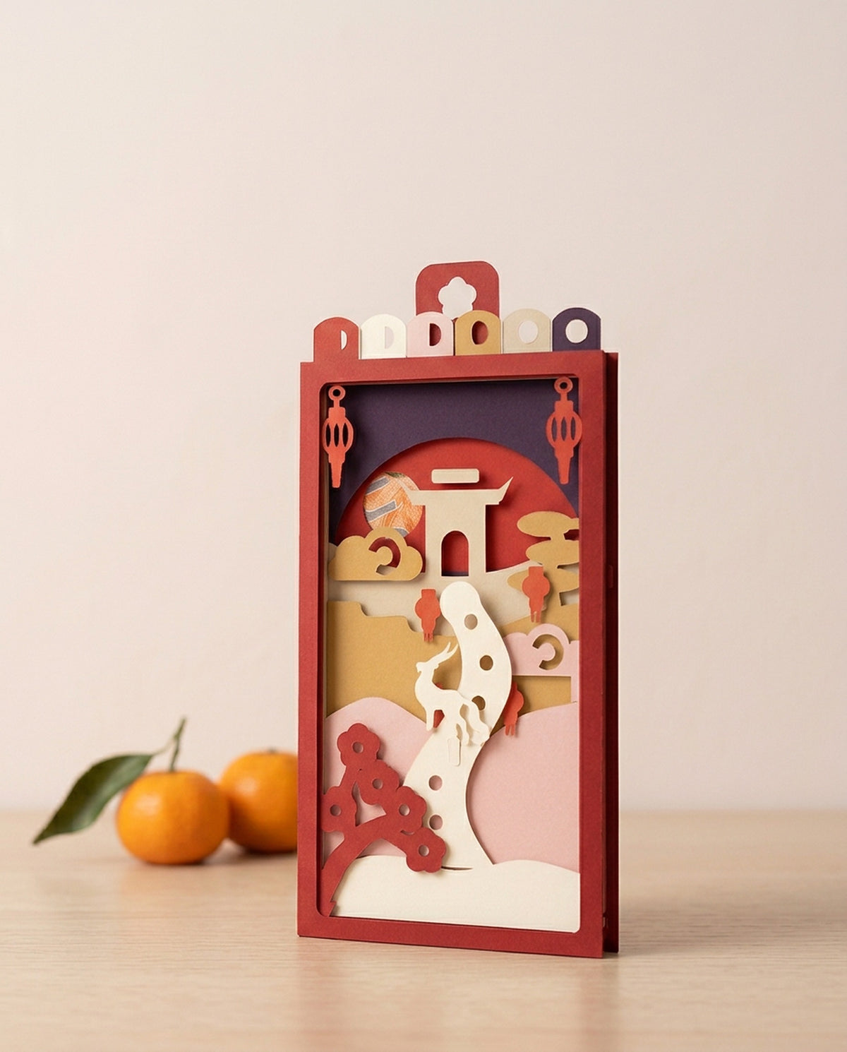 Year of the Goat Pocket Story, a paper-art hóngbāo for Chinese New Year, standing on a table with mandarins